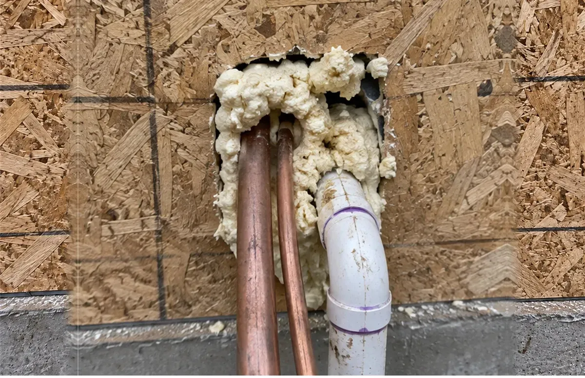 Copper lines and PVC drain through a hole in sheathing packed with expanding foam